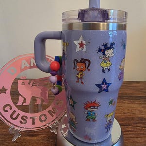 Puede incluir: Un vaso con purpurina lavanda con asa, con calcoman&iacute;as de personajes de dibujos animados y detalles de estrellas. El vaso tiene una tapa transparente y una tapa abatible morada. Un dije de cuentas est&aacute; unido al asa.