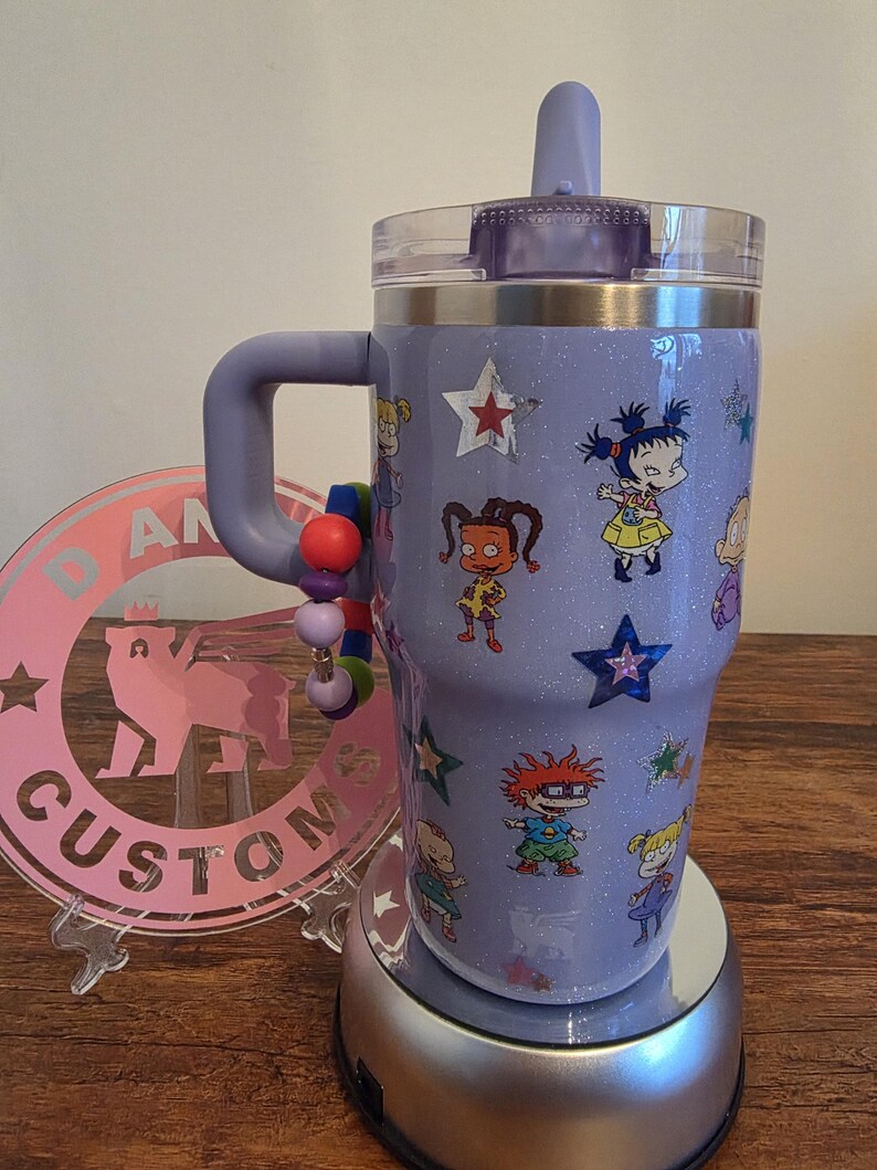 Puede incluir: Un vaso con purpurina lavanda con asa, con calcoman&iacute;as de personajes de dibujos animados y detalles de estrellas. El vaso tiene una tapa transparente y una tapa abatible morada. Un dije de cuentas est&aacute; unido al asa.