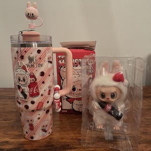 Pu&ograve; includere: Un bicchiere Stanley rosa e bianco con un design a coniglietto, un topper per cannuccia a coniglietto abbinato e una statuina a coniglietto che tiene in mano una bottiglia di Coca-Cola. &Egrave; visibile anche una scatola con un design a mostro.