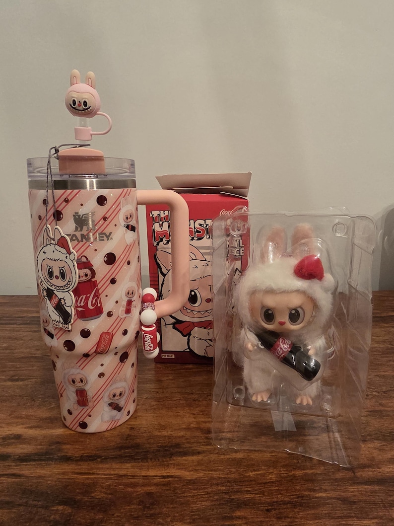 Pu&ograve; includere: Un bicchiere Stanley rosa e bianco con un design a coniglietto, un topper per cannuccia a coniglietto abbinato e una statuina a coniglietto che tiene in mano una bottiglia di Coca-Cola. &Egrave; visibile anche una scatola con un design a mostro.