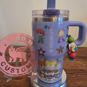 Puede incluir: Un vaso Stanley lavanda con personajes de dibujos animados de Rugrats y estrellas. El vaso tiene un asa, una tapa transparente y una pajita morada. Un charm con cuentas de colores cuelga del asa. El vaso est&aacute; sobre una base plateada.