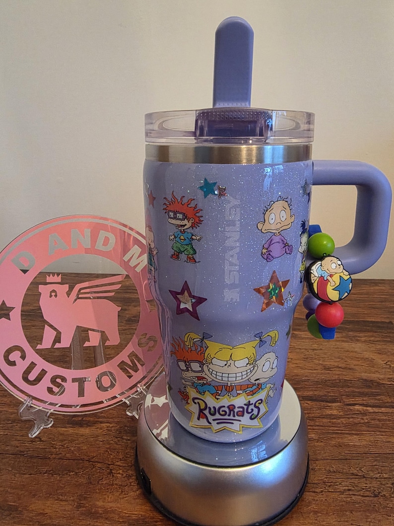 Puede incluir: Un vaso Stanley lavanda con personajes de dibujos animados de Rugrats y estrellas. El vaso tiene un asa, una tapa transparente y una pajita morada. Un charm con cuentas de colores cuelga del asa. El vaso est&aacute; sobre una base plateada.