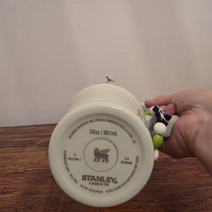 Puede incluir: Un mug Stanley blanco con un asa decorativa. El mug est&aacute; marcado como 887 ml, apto para lavavajillas y sin BPA. El asa tiene cuentas de colores y una peque&ntilde;a figura de p&aacute;jaro.