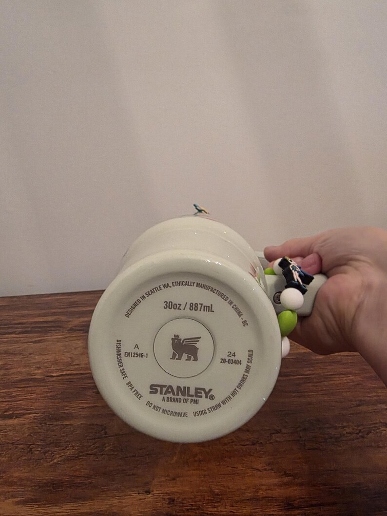 Puede incluir: Un mug Stanley blanco con un asa decorativa. El mug est&aacute; marcado como 887 ml, apto para lavavajillas y sin BPA. El asa tiene cuentas de colores y una peque&ntilde;a figura de p&aacute;jaro.