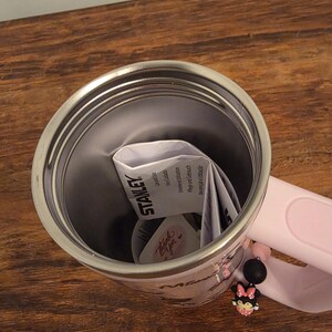 May include: A stainless steel Stanley tumbler with a pink handle and a Minnie Mouse design. The tumbler has a pink strap with a Minnie Mouse charm. The interior of the tumbler is silver, and the handle is pink.