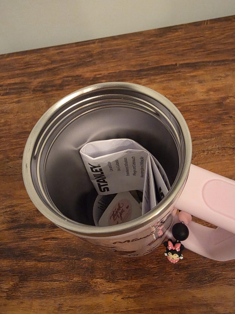 May include: A stainless steel Stanley tumbler with a pink handle and a Minnie Mouse design. The tumbler has a pink strap with a Minnie Mouse charm. The interior of the tumbler is silver, and the handle is pink.