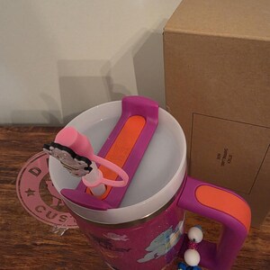 May include: A pink glittery travel mug with a purple handle and lid. The lid has an orange slider and a pink butterfly charm. A charm with white beads and a blue cartoon character hangs from the handle. A cardboard box is in the background.