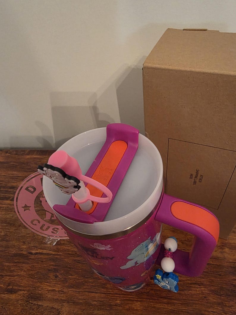 May include: A pink glittery travel mug with a purple handle and lid. The lid has an orange slider and a pink butterfly charm. A charm with white beads and a blue cartoon character hangs from the handle. A cardboard box is in the background.