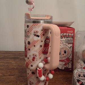 Pu&ograve; includere: Un bicchiere rosa e bianco con manico, con un design a coniglietto dei cartoni animati e il marchio Coca-Cola. Il bicchiere ha un coperchio rosa abbinato e un topper per cannuccia a forma di coniglio. Un braccialetto con ciondoli di perline &egrave; attaccato al manico.