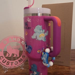 May include: A pink glitter Stanley tumbler with Care Bear designs. The tumbler has a white lid, a purple handle with an orange accent, and a decorative charm. A pink and white logo is in the background.
