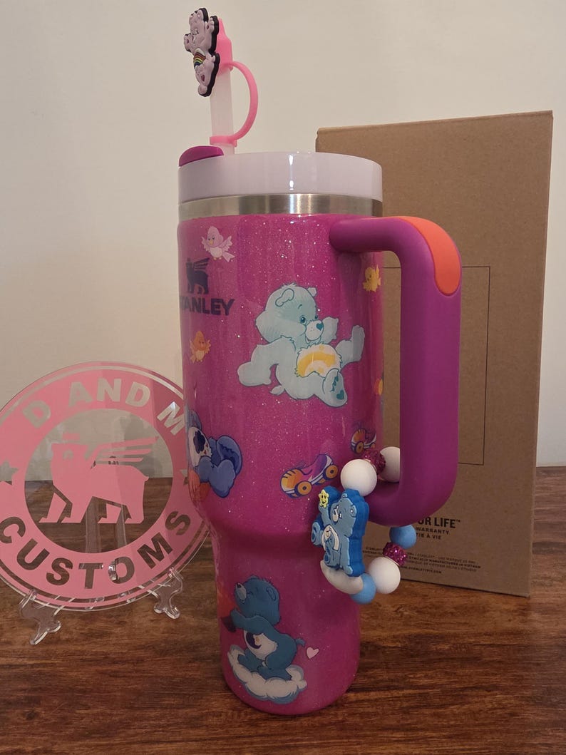 May include: A pink glitter Stanley tumbler with Care Bear designs. The tumbler has a white lid, a purple handle with an orange accent, and a decorative charm. A pink and white logo is in the background.