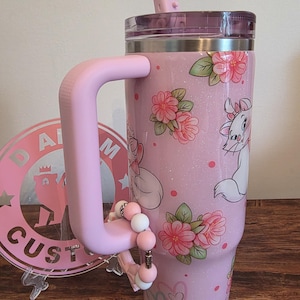 May include: A light pink glitter tumbler with a handle and a straw, featuring a repeating pattern of white cartoon cats with pink bows, pink floral clusters, and small pink hearts. A beaded charm is attached to the handle of the drink container.