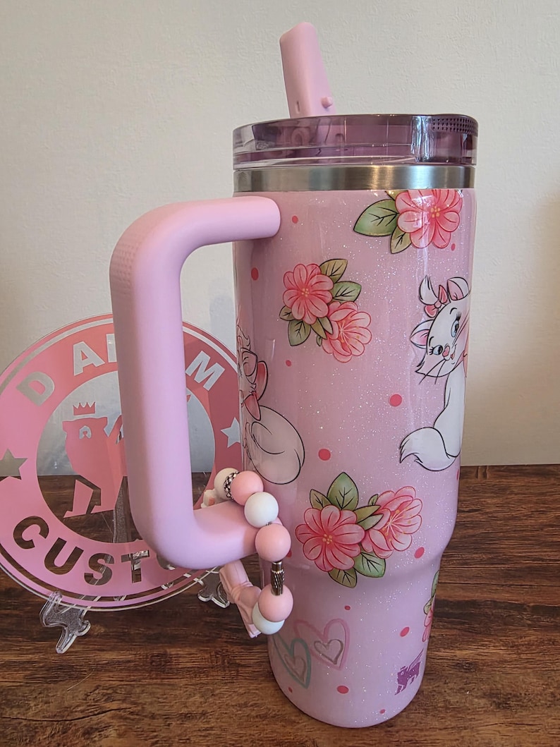 May include: A light pink glitter tumbler with a handle and a straw, featuring a repeating pattern of white cartoon cats with pink bows, pink floral clusters, and small pink hearts. A beaded charm is attached to the handle of the drink container.