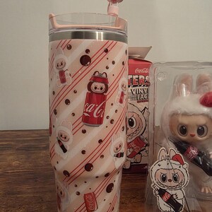 Pu&ograve; includere: Un bicchiere rosa e bianco con coperchio e cannuccia, con un motivo ripetuto di coniglietti dei cartoni animati e lattine di Coca-Cola. Il design include strisce diagonali rosse e pois marroni. Sono visibili anche una scatola di Coca-Cola e una statuina di coniglio.