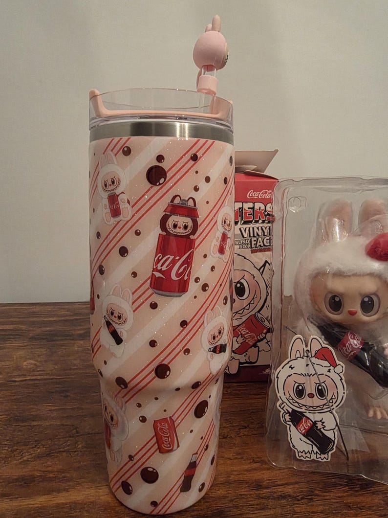 Pu&ograve; includere: Un bicchiere rosa e bianco con coperchio e cannuccia, con un motivo ripetuto di coniglietti dei cartoni animati e lattine di Coca-Cola. Il design include strisce diagonali rosse e pois marroni. Sono visibili anche una scatola di Coca-Cola e una statuina di coniglio.