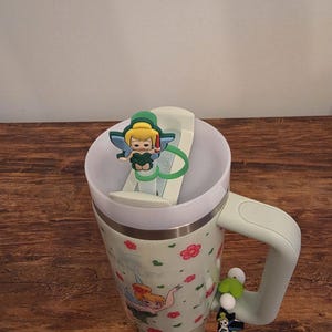 Puede incluir: Un termo de viaje verde menta con tapa y asa blancas. El termo presenta un estampado floral y una imagen de dibujos animados de Campanilla. Un dije de Campanilla est&aacute; sujeto al asa y otro a la tapa.