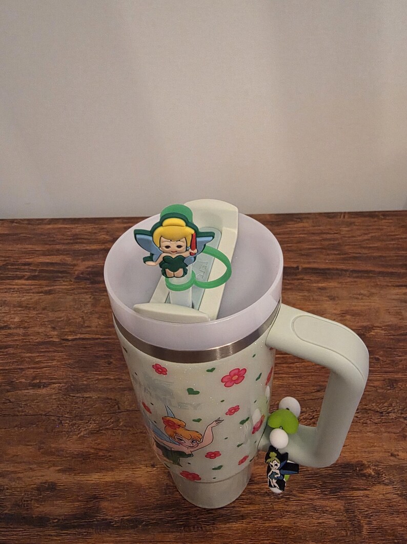 Puede incluir: Un termo de viaje verde menta con tapa y asa blancas. El termo presenta un estampado floral y una imagen de dibujos animados de Campanilla. Un dije de Campanilla est&aacute; sujeto al asa y otro a la tapa.