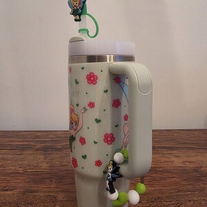 Puede incluir: Un vaso verde claro con asa, decorado con flores rosas, corazones verdes y un hada de dibujos animados. El vaso tiene una tapa blanca y una pajita verde con un adorno de hada. Un adorno de cuentas con un hada est&aacute; unido al asa.