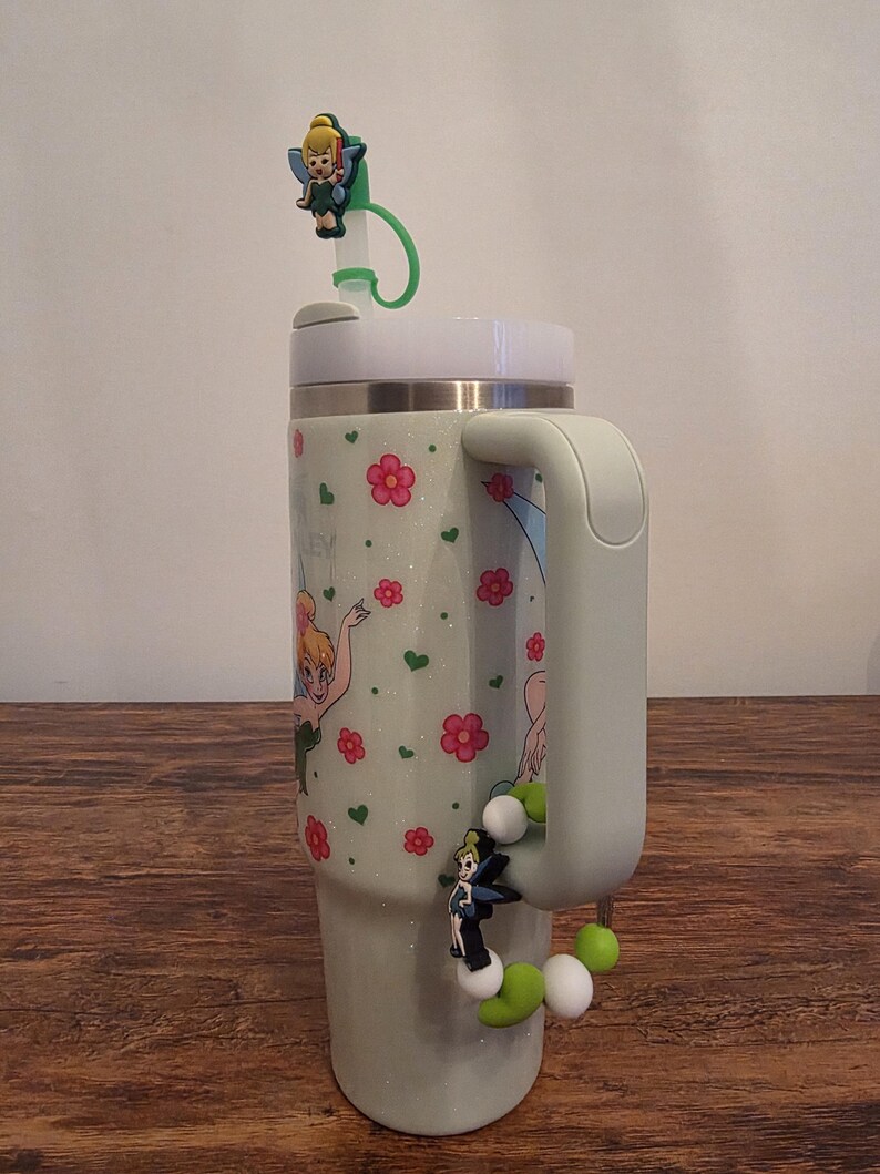 Puede incluir: Un vaso verde claro con asa, decorado con flores rosas, corazones verdes y un hada de dibujos animados. El vaso tiene una tapa blanca y una pajita verde con un adorno de hada. Un adorno de cuentas con un hada est&aacute; unido al asa.