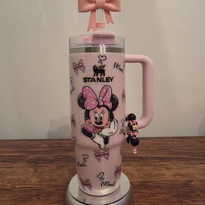 May include: A pink Stanley tumbler featuring Minnie Mouse. The tumbler has a pink bow on top and a handle. The design includes Minnie Mouse, bows, and the word "Minnie". A charm of Minnie Mouse is attached to the handle.