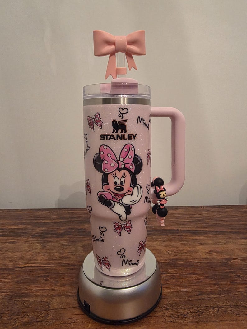 May include: A pink Stanley tumbler featuring Minnie Mouse. The tumbler has a pink bow on top and a handle. The design includes Minnie Mouse, bows, and the word "Minnie". A charm of Minnie Mouse is attached to the handle.