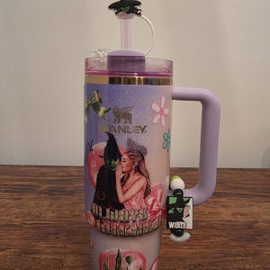 May include: A large, insulated tumbler with a purple handle and lid, featuring a design from the musical "Wicked." The tumbler has a straw and a decorative charm. The design includes images of characters and the word "Wicked."