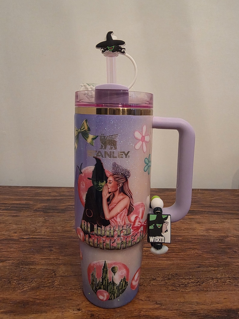 May include: A large, insulated tumbler with a purple handle and lid, featuring a design from the musical "Wicked." The tumbler has a straw and a decorative charm. The design includes images of characters and the word "Wicked."