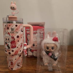 Pu&ograve; includere: Un bicchiere Stanley rosa con un design a coniglietto, un manico abbinato e un tappo per cannuccia a forma di coniglietto. Una piccola figura di coniglietto di peluche bianco con un fiocco rosso e una bottiglia di Coca-Cola.