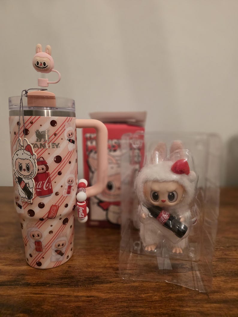 Pu&ograve; includere: Un bicchiere Stanley rosa con un design a coniglietto, un manico abbinato e un tappo per cannuccia a forma di coniglietto. Una piccola figura di coniglietto di peluche bianco con un fiocco rosso e una bottiglia di Coca-Cola.