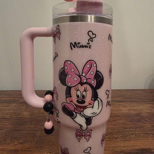 May include: Pink glitter tumbler featuring Minnie Mouse with a polka dot bow. The tumbler has a pink handle and a pink bow on the lid. The word "Minnie" is written on the tumbler. The tumbler has a capacity of 30 ounces.