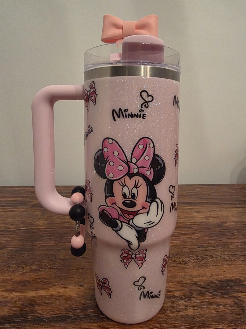 May include: Pink glitter tumbler featuring Minnie Mouse with a polka dot bow. The tumbler has a pink handle and a pink bow on the lid. The word "Minnie" is written on the tumbler. The tumbler has a capacity of 30 ounces.