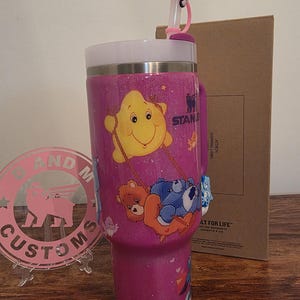 May include: A pink glittery Stanley tumbler featuring Care Bears characters. The tumbler has a white rim and handle, with a pink straw and a decorative topper. The tumbler is decorated with various Care Bears characters.