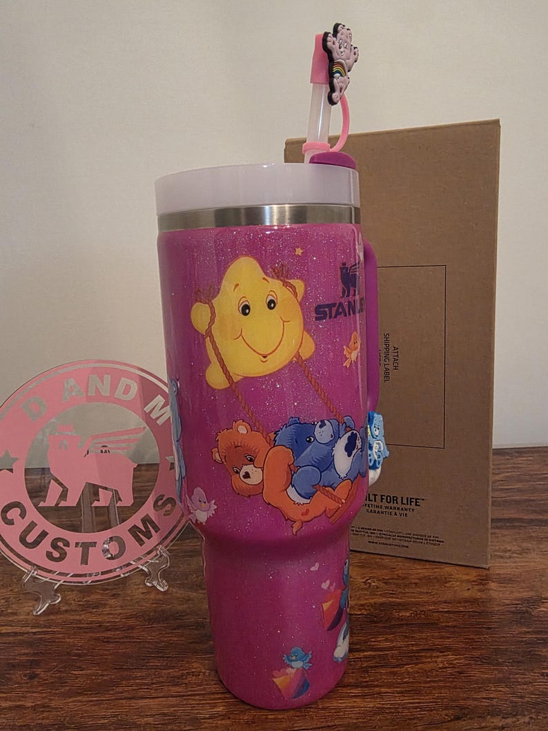 May include: A pink glittery Stanley tumbler featuring Care Bears characters. The tumbler has a white rim and handle, with a pink straw and a decorative topper. The tumbler is decorated with various Care Bears characters.