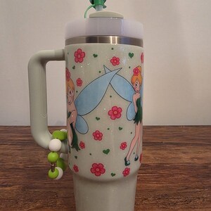 Puede incluir: Un vaso verde claro con asa, con un dise&ntilde;o de hada con alas azules y flores rosas. El vaso tiene una tapa blanca y una pajita verde. Un charm de cuentas cuelga del asa.