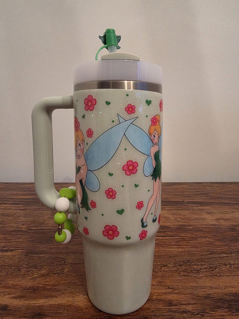 Puede incluir: Un vaso verde claro con asa, con un dise&ntilde;o de hada con alas azules y flores rosas. El vaso tiene una tapa blanca y una pajita verde. Un charm de cuentas cuelga del asa.