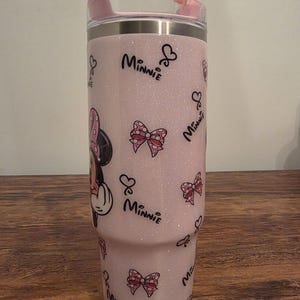 May include: A pink glitter tumbler featuring Minnie Mouse. The tumbler has a pink lid and straw. The design includes Minnie Mouse, bows, and the word "Minnie" in a script font. The tumbler is designed to hold approximately 30 ounces.