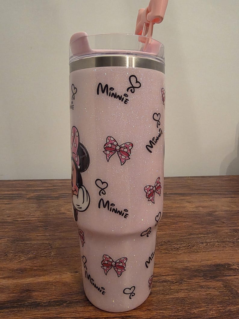 May include: A pink glitter tumbler featuring Minnie Mouse. The tumbler has a pink lid and straw. The design includes Minnie Mouse, bows, and the word "Minnie" in a script font. The tumbler is designed to hold approximately 30 ounces.
