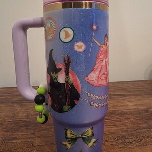 May include: A large, insulated tumbler with a purple handle and lid. The tumbler is decorated with images from The Wizard of Oz, including the Wicked Witch, a fairy, and a bow. The background is a sparkly blue.