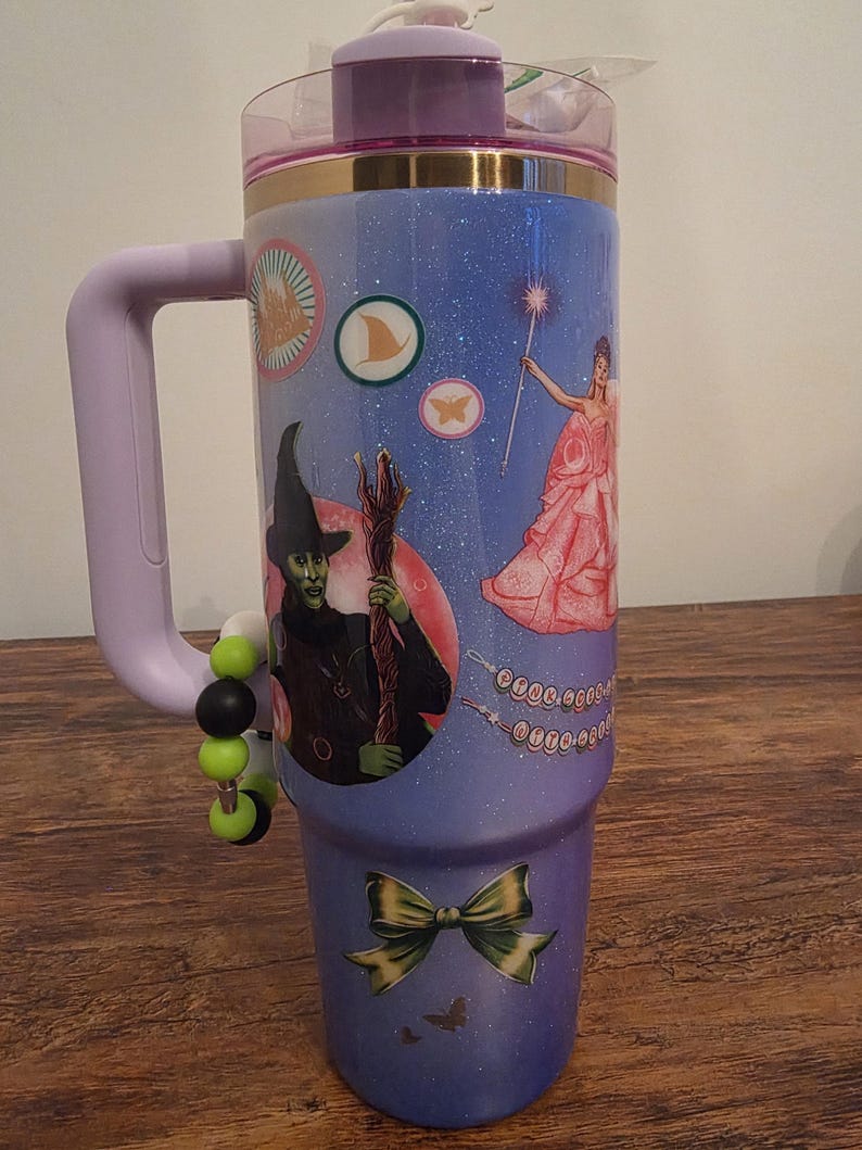 May include: A large, insulated tumbler with a purple handle and lid. The tumbler is decorated with images from The Wizard of Oz, including the Wicked Witch, a fairy, and a bow. The background is a sparkly blue.