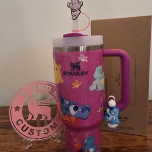 May include: A pink glitter Stanley tumbler featuring Care Bears characters. The tumbler has a purple handle and a matching straw with a Care Bear topper. A pink custom logo is in the foreground.