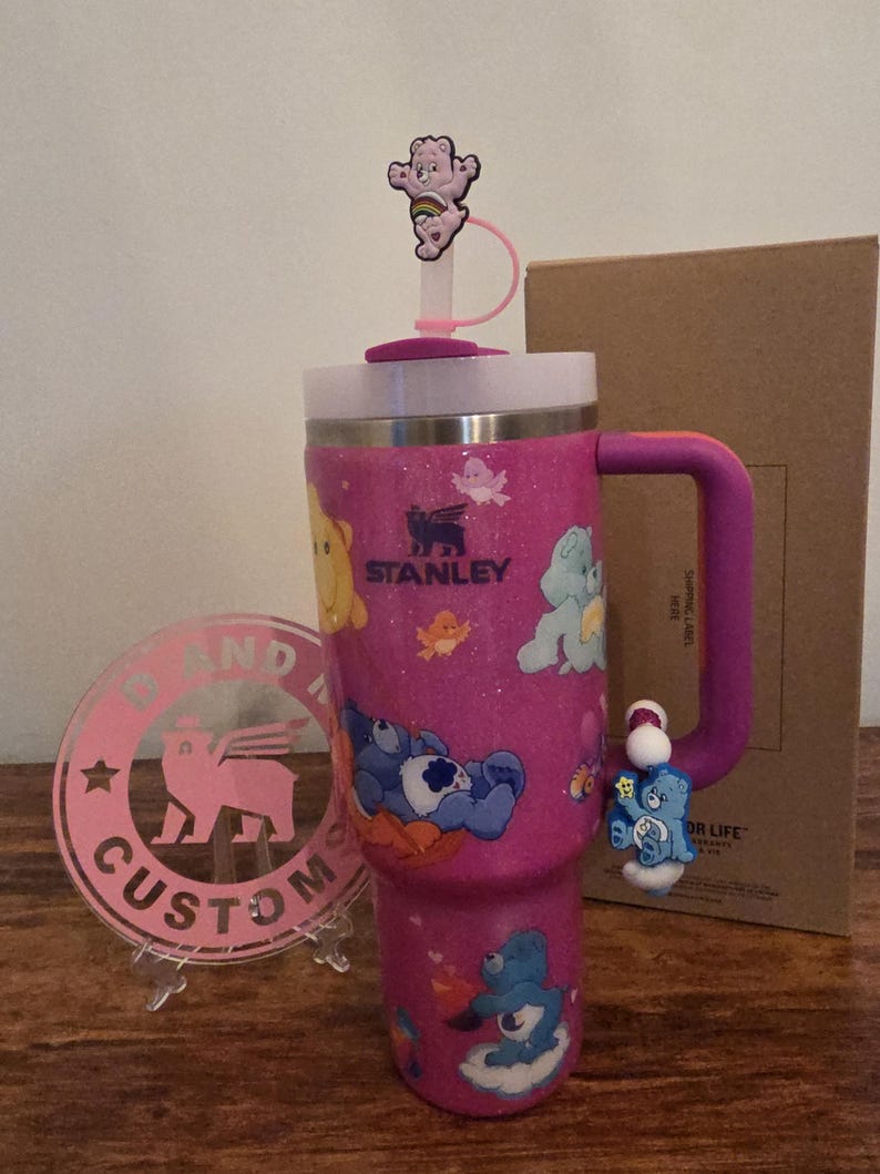 May include: A pink glitter Stanley tumbler featuring Care Bears characters. The tumbler has a purple handle and a matching straw with a Care Bear topper. A pink custom logo is in the foreground.