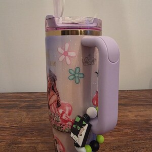 May include: A large, insulated tumbler with a lavender handle and lid, decorated with floral and princess-themed graphics. The tumbler features a straw and a charm of a witch on top. A charm with the word "Wicked" is attached to the handle.