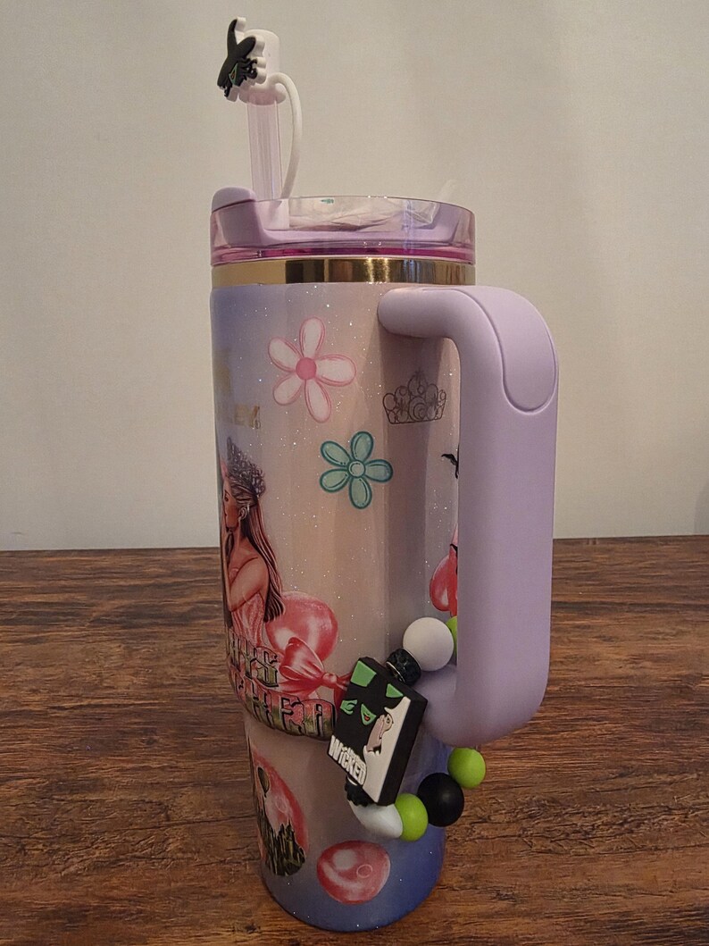 May include: A large, insulated tumbler with a lavender handle and lid, decorated with floral and princess-themed graphics. The tumbler features a straw and a charm of a witch on top. A charm with the word "Wicked" is attached to the handle.