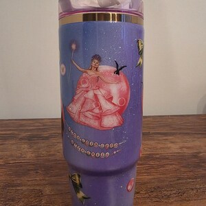 May include: A purple tumbler with a fairy design. The tumbler features a princess in a pink dress with a wand, surrounded by stars and decorative bows. The lid is pink with a gold band.