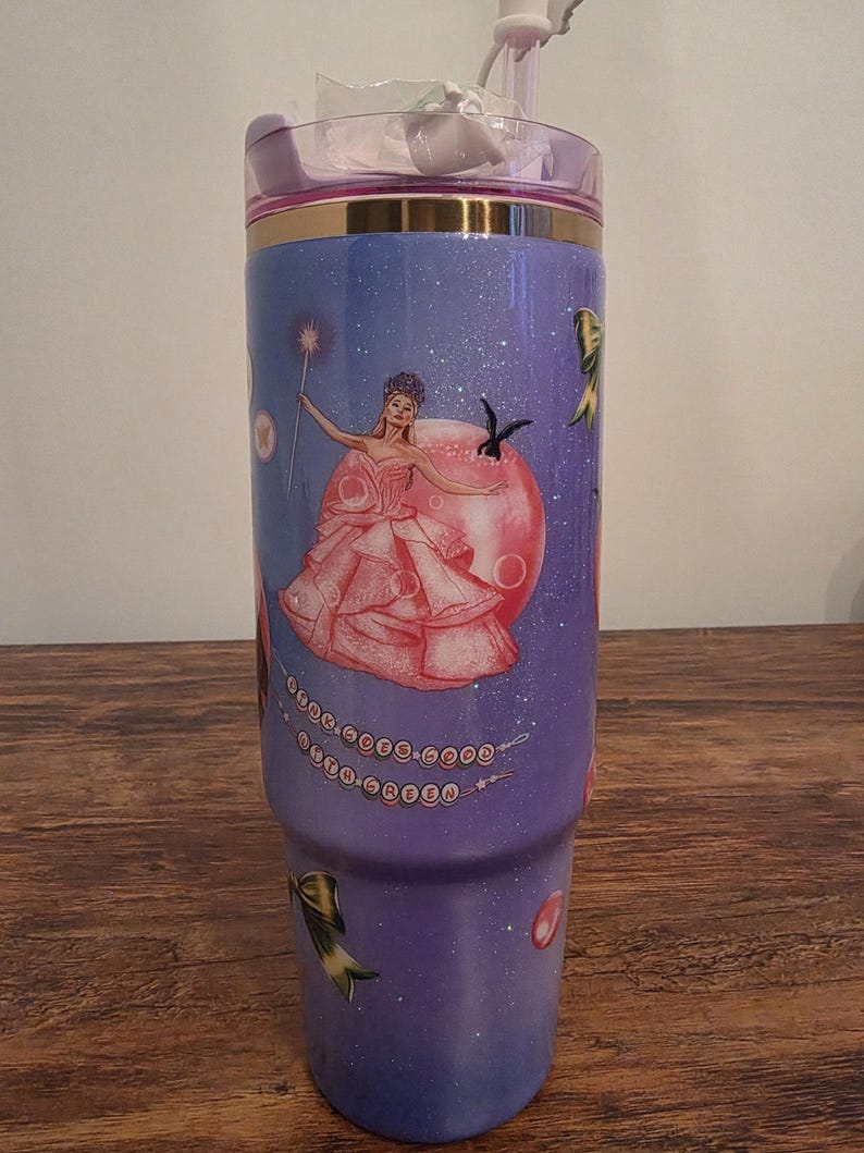 May include: A purple tumbler with a fairy design. The tumbler features a princess in a pink dress with a wand, surrounded by stars and decorative bows. The lid is pink with a gold band.