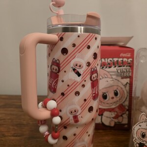 Pu&ograve; includere: Un bicchiere rosa e bianco con manico, con un design a tema coniglietto con marchio Coca-Cola. Il bicchiere ha un coperchio rosa e un braccialetto di perline decorativo. Il design include strisce rosse, pois e personaggi dei cartoni animati.