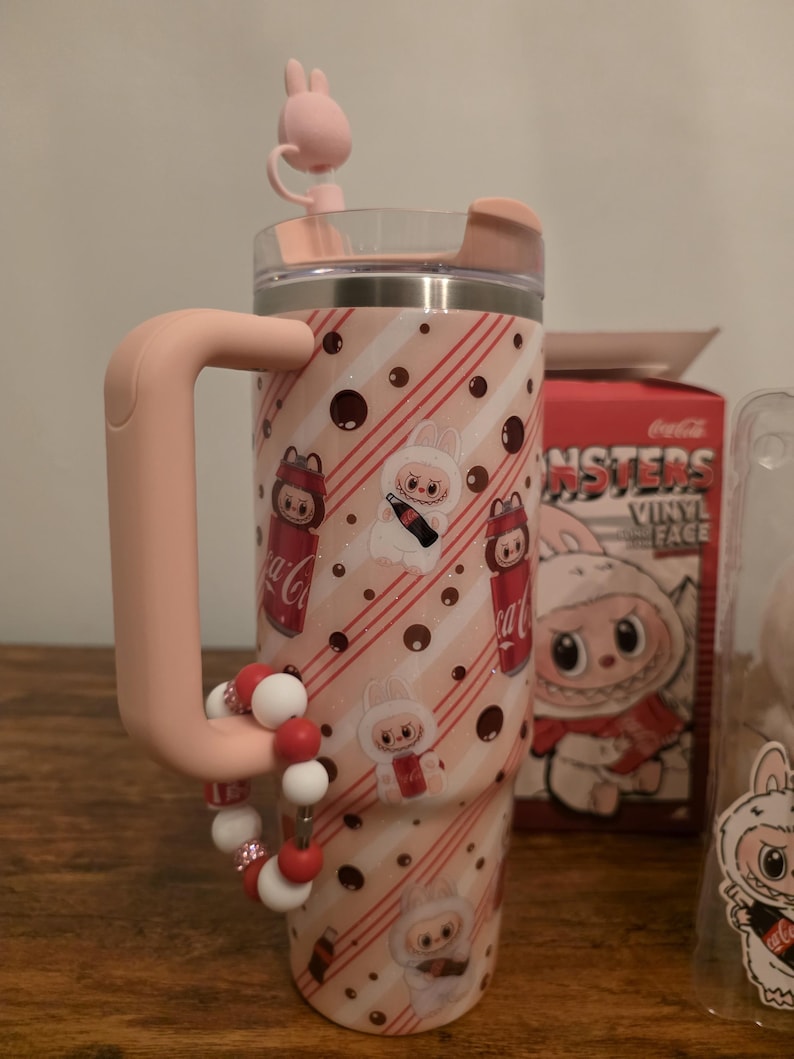 Pu&ograve; includere: Un bicchiere rosa e bianco con manico, con un design a tema coniglietto con marchio Coca-Cola. Il bicchiere ha un coperchio rosa e un braccialetto di perline decorativo. Il design include strisce rosse, pois e personaggi dei cartoni animati.