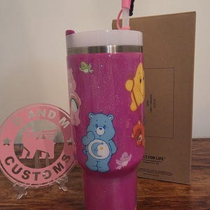 May include: A pink glitter tumbler featuring Care Bears characters. The tumbler has a white lid, a pink straw, and a clear plastic base. The design includes a blue bear with a crescent moon, a yellow bear, and other Care Bears characters.