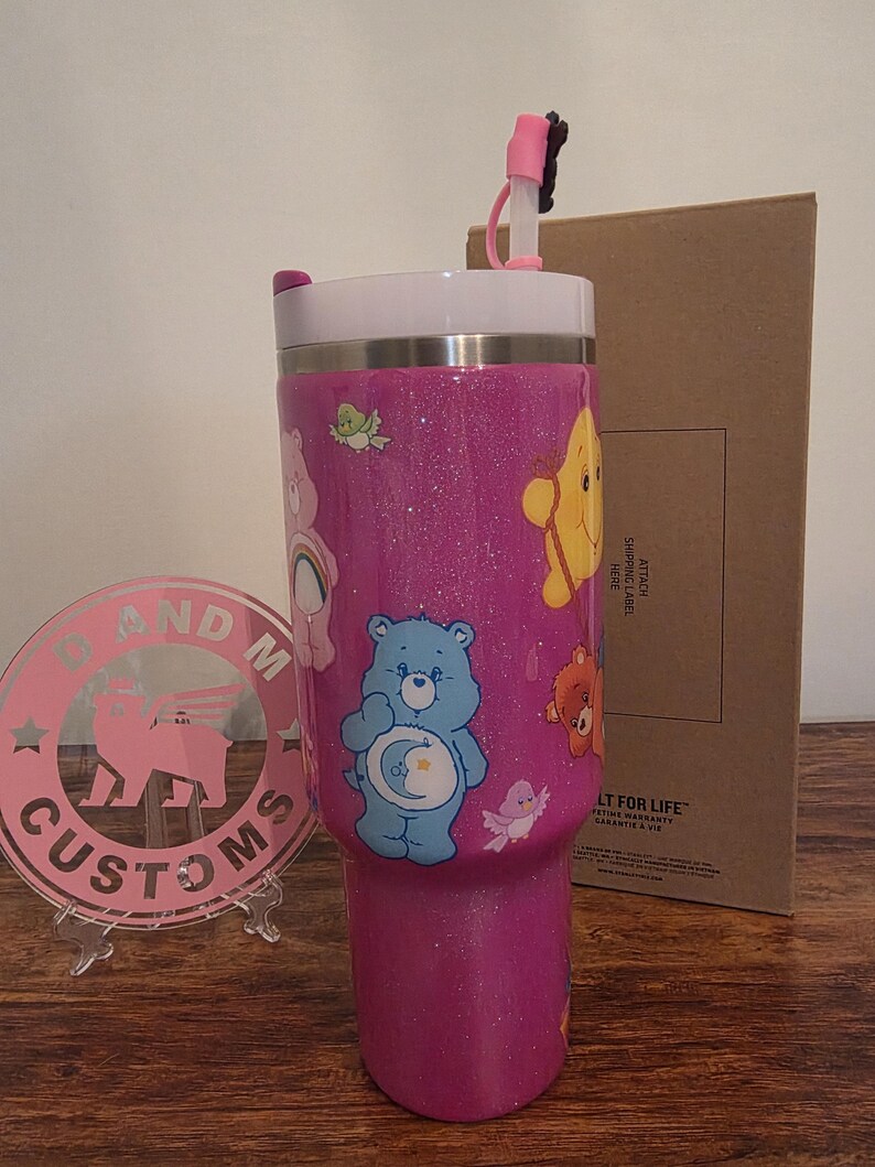May include: A pink glitter tumbler featuring Care Bears characters. The tumbler has a white lid, a pink straw, and a clear plastic base. The design includes a blue bear with a crescent moon, a yellow bear, and other Care Bears characters.