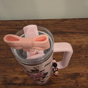 May include: Pink Stanley tumbler with a Minnie Mouse design. The tumbler has a clear lid, a pink handle, and a pink bow-shaped lid closure. A Minnie Mouse charm hangs from the handle. The tumbler is on a wooden surface.